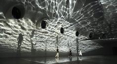 some people are walking around in a large room with light streaming through the ceiling and shadows on the walls