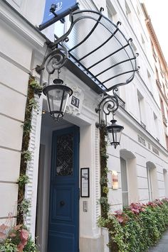 a blue door is in front of a white building with flowers on the side and an awning over it