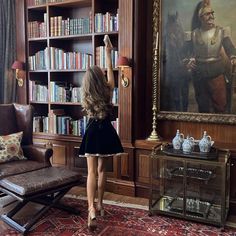 a woman standing in front of a book shelf with books on it and a painting behind her