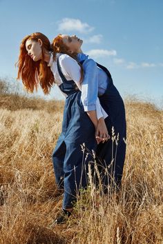 two women in overalls are standing in tall grass and one is holding the other's head