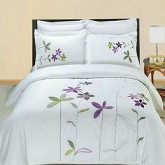 a white bed with purple and green flowers on the comforter, next to a night stand