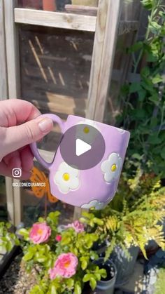 someone is holding a purple mug with white flowers on it in front of some potted plants