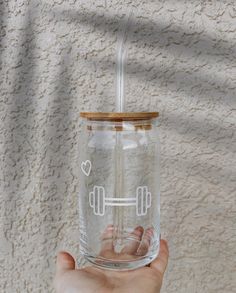 a hand holding a clear glass with a wooden lid and straw in front of a white wall