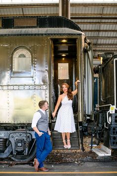 a man standing next to a woman in front of an old train car holding hands