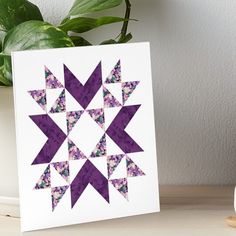 a purple and white quilt art board on a table next to a potted plant