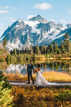 a bride and groom standing on a dock in front of a mountain lake holding hands