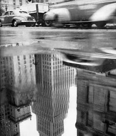 a black and white photo of a city street with buildings reflected in the wet water