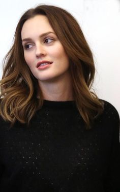 a woman with long brown hair wearing a black sweater and standing in front of a white wall