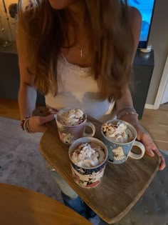 a woman holding two cups of hot chocolate and whipped cream on top of a wooden tray