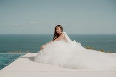 a woman in a wedding dress sitting on the edge of a pool by the ocean