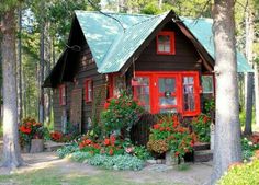 a small cabin in the woods with red doors and flowers on the front porch, surrounded by trees