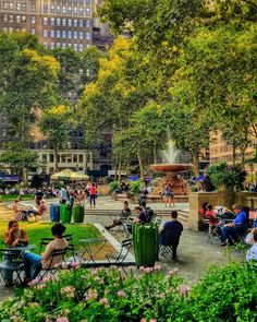 people are sitting at tables in the park with flowers and trees around them, surrounded by tall buildings