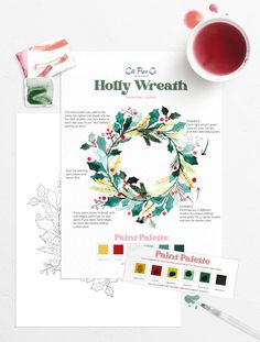 the holly wreath pattern is shown next to a cup of tea and watercolor pencils