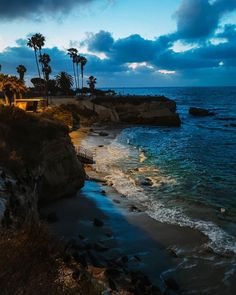 the beach is lined with palm trees and rocky cliffs at dusk, along with dark clouds in the sky