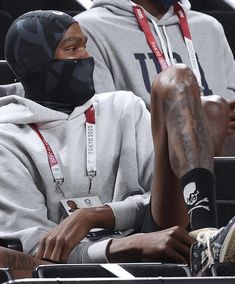 two men sitting next to each other at a basketball game wearing face coverings and hoodies