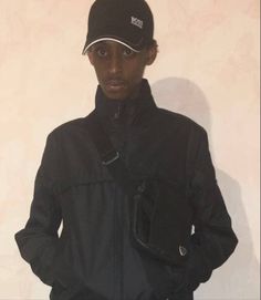 a young man wearing a black jacket and hat standing in front of a white wall