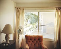 an orange chair sitting in front of a window next to a table with a fan