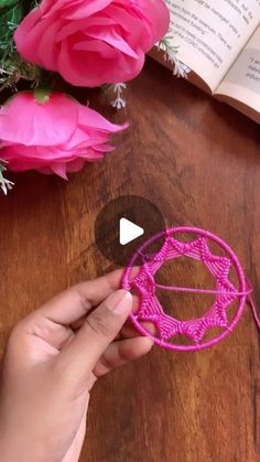 someone is making a crochet flower decoration with pink yarn and flowers in the background