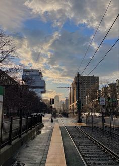 an empty train track next to a street with buildings on both sides and clouds in the sky