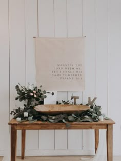 a wooden table topped with greenery next to a white wall and a framed sign