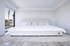 a large white bed sitting inside of a bedroom next to a tall glass window on top of a hard wood floor