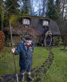 an old man holding a stick in front of a hobbot house on the side of a road