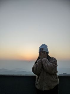 a person wearing a hat is looking at the sunset with their hands to their face