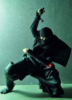 a man dressed in black is holding a knife up to his head while standing on one leg