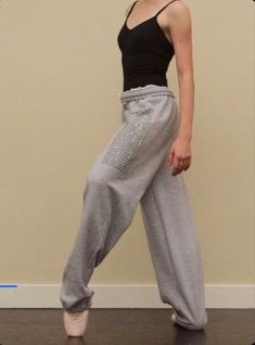 a woman standing in front of a wall with her legs crossed and wearing grey sweatpants