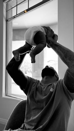 a man holding a baby up to his head while sitting in front of a window