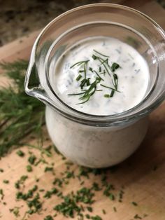 a jar filled with white sauce sitting on top of a wooden table next to green herbs