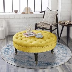 a yellow ottoman sits on a blue rug in front of a white brick wall and window