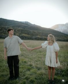 a man and woman holding hands while standing in a field with mountains in the background