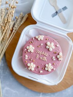 a pink frosted cake with white flowers on it sitting in a plastic container next to some wheat stalks