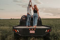 two people standing on the back of a pickup truck in a grassy field at sunset