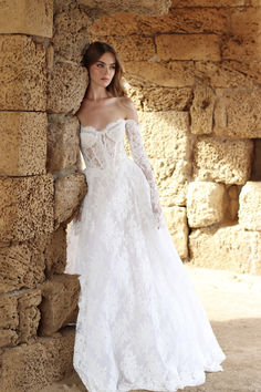 a woman in a white wedding dress leaning against a stone wall with her hands on her hips