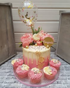 there is a cake with pink frosting and flowers on the top, surrounded by cupcakes