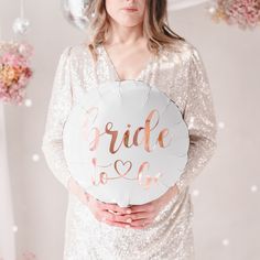 a woman in a white dress holding a bride to be balloon with rose gold foil lettering