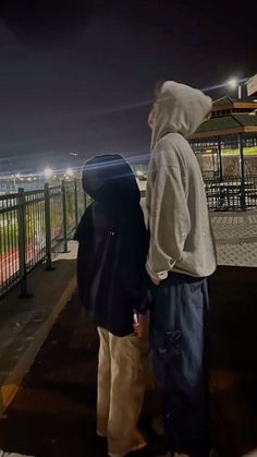 two people standing next to each other on a sidewalk at night with the city lights in the background