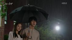 a man and woman standing under an umbrella in the rain with lights on behind them