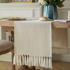 a white table cloth with tassels on it and a blue vase filled with flowers in the background