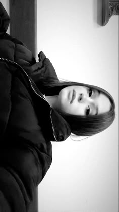 black and white photograph of a woman in a coat looking at the camera while standing next to a wall