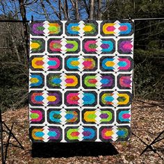 a colorful quilt hanging on a clothesline in the woods with trees and leaves around it