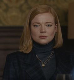 a woman with blonde hair wearing a black turtle neck sweater and gold necklace, looking at the camera