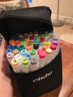 a bag filled with markers and pens on top of a wooden table next to a person's hand