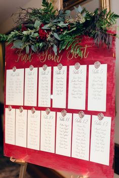 a red and white wedding seating chart with greenery on the top is displayed in front of a mirror