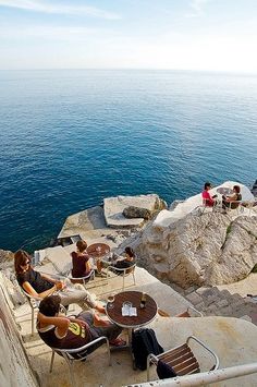 people sitting on the edge of a cliff overlooking the ocean with chairs and tables in front of them