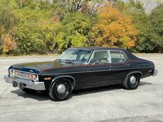 an old black car is parked in a parking lot with trees and bushes behind it