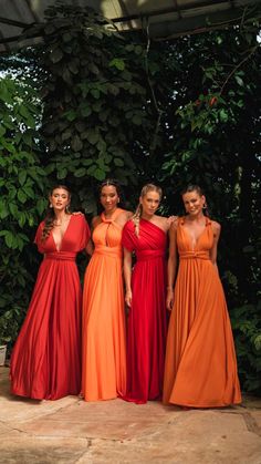 four women in long dresses standing next to each other on a stone floor with greenery behind them