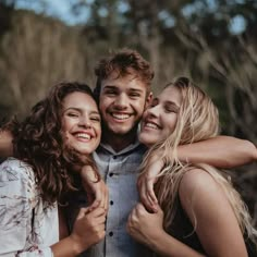 three young people are hugging and smiling for the camera while standing in front of some trees